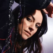 Portrait of Scottish singer-songwriter Amy Macdonald in a close-up shot. She is leaning slightly forward with her arm resting above her head, gazing directly at the camera. The background is softly lit in purples and greys, and strands of beaded fabric add texture to the foreground.
