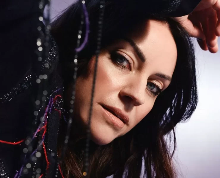 Portrait of Scottish singer-songwriter Amy Macdonald in a close-up shot. She is leaning slightly forward with her arm resting above her head, gazing directly at the camera. The background is softly lit in purples and greys, and strands of beaded fabric add texture to the foreground.