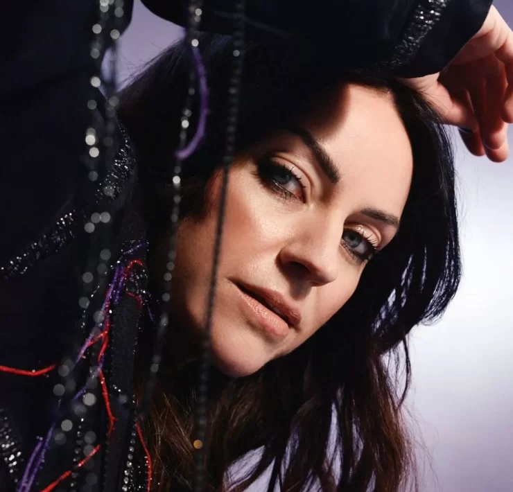 Portrait of Scottish singer-songwriter Amy Macdonald in a close-up shot. She is leaning slightly forward with her arm resting above her head, gazing directly at the camera. The background is softly lit in purples and greys, and strands of beaded fabric add texture to the foreground.