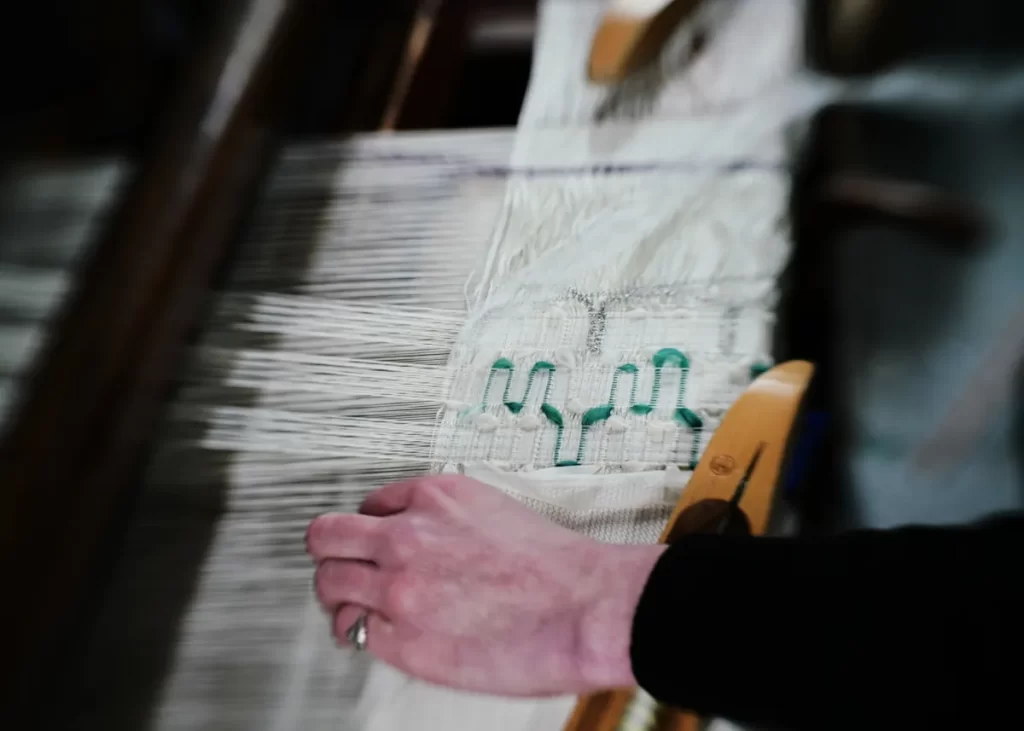 Close-up of hands weaving a white fabric with green detailing on a loom