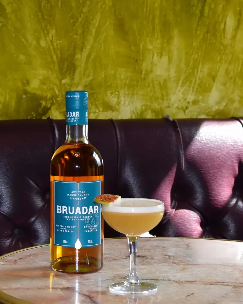 Bruadar whisky bottle and The Hot Honey cocktail on a marble table in Kelvingrove Café, Glasgow, set against a rich olive green and burgundy background.