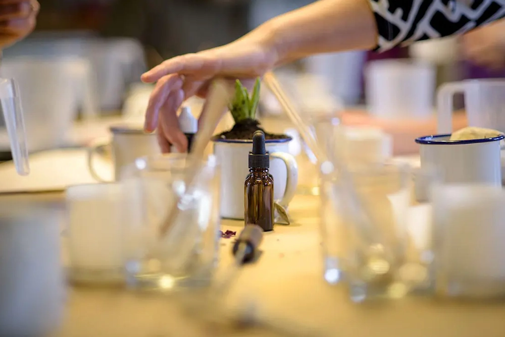 A hand reaches across a workshop table with oils, jars, and tools arranged for candle making.