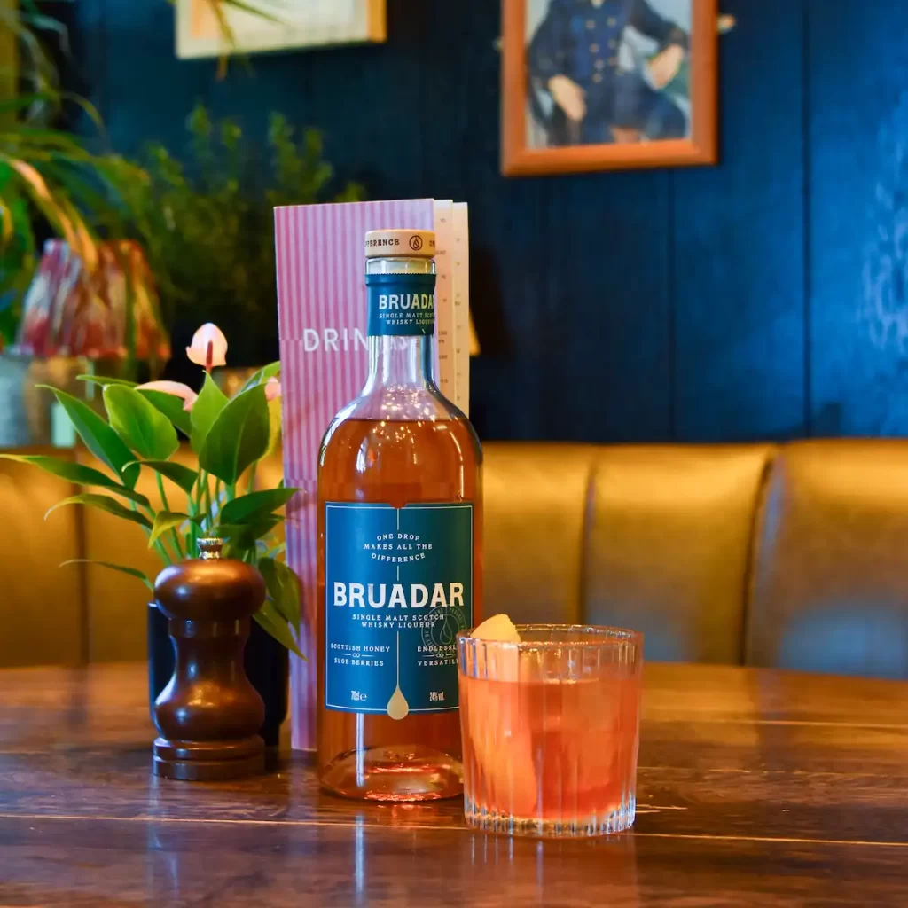 Bruadar whisky bottle and Bee-yond the Smoke cocktail on a rustic wooden table at Montpeliers in Edinburgh, surrounded by plants and warm-toned interiors.