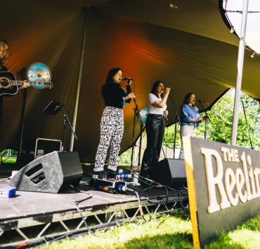 Folk musicians perform onstage at The Reeling Festival in Glasgow, under a canopy tent at Rouken Glen Park.