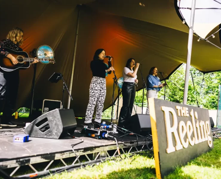 Folk musicians perform onstage at The Reeling Festival in Glasgow, under a canopy tent at Rouken Glen Park.