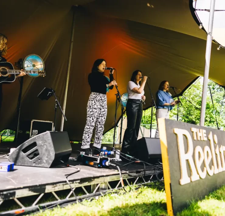 Folk musicians perform onstage at The Reeling Festival in Glasgow, under a canopy tent at Rouken Glen Park.