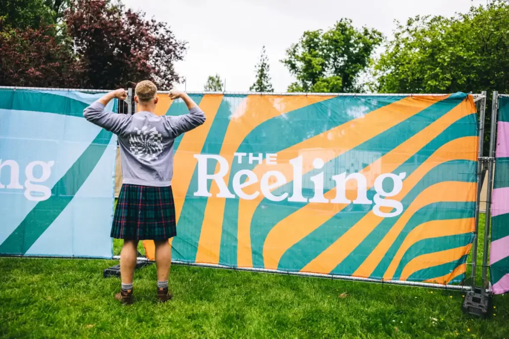 A man in a kilt poses in front of a colourful Reeling Festival banner at Rouken Glen Park in Glasgow.