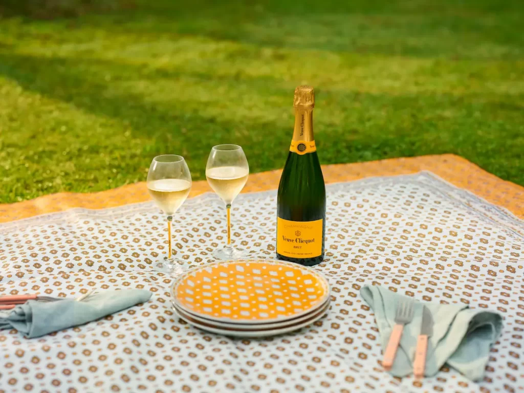 A Veuve Clicquot Champagne bottle and two glasses set on a patterned picnic blanket with yellow plates and napkins, laid out on a manicured lawn.