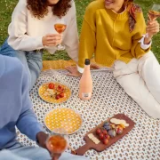 Three people enjoying rosé wine and a fresh picnic with yellow plates, a grazing board, and a 'Let's Picnic' bottle on a patterned blanket outdoors.