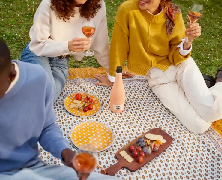 Three people enjoying rosé wine and a fresh picnic with yellow plates, a grazing board, and a 'Let's Picnic' bottle on a patterned blanket outdoors.