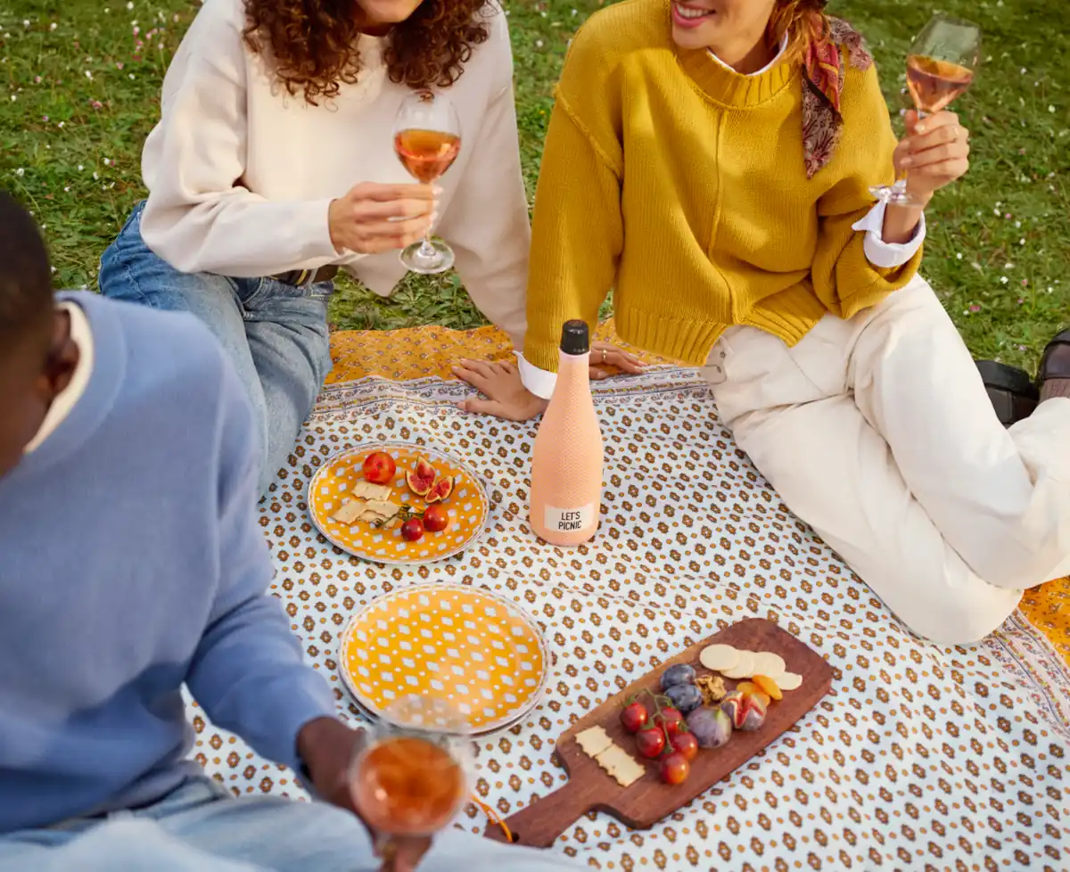 Three people enjoying rosé wine and a fresh picnic with yellow plates, a grazing board, and a 'Let's Picnic' bottle on a patterned blanket outdoors.