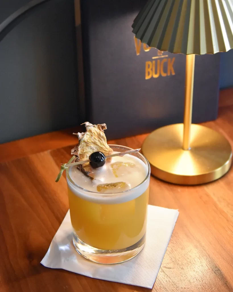 The Pineapple Swizzle cocktail served on a wooden table at The Voyage of Buck, garnished with dried pineapple and cherry, next to a gold lamp and cocktail menu.