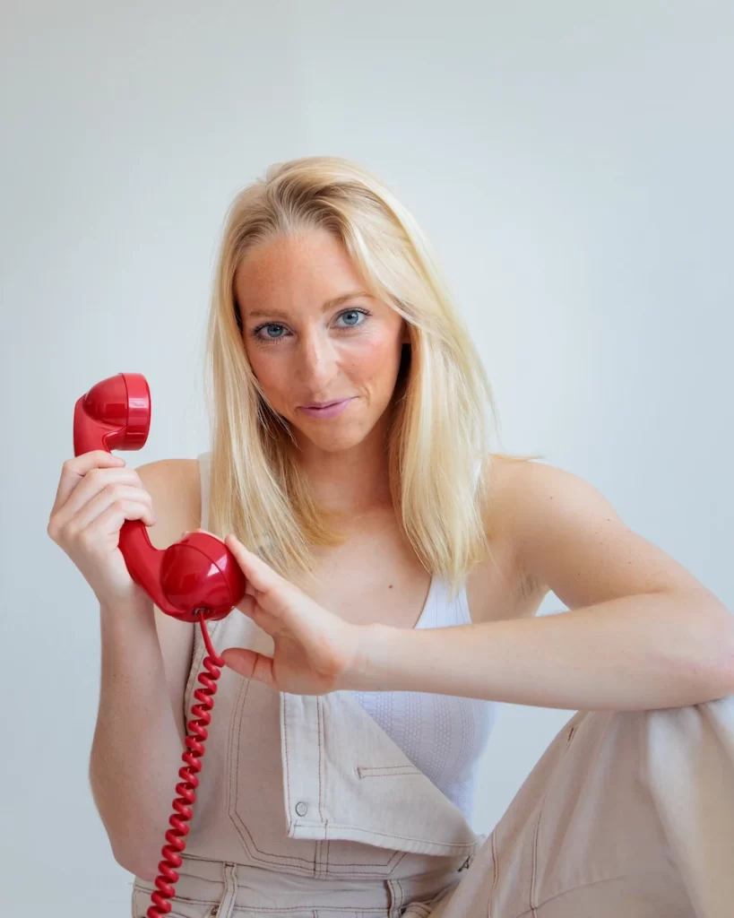 Close-up of Olivia Raine Atwood smiling with a red telephone in her hand, wearing cream dungarees and white vest, on a white background