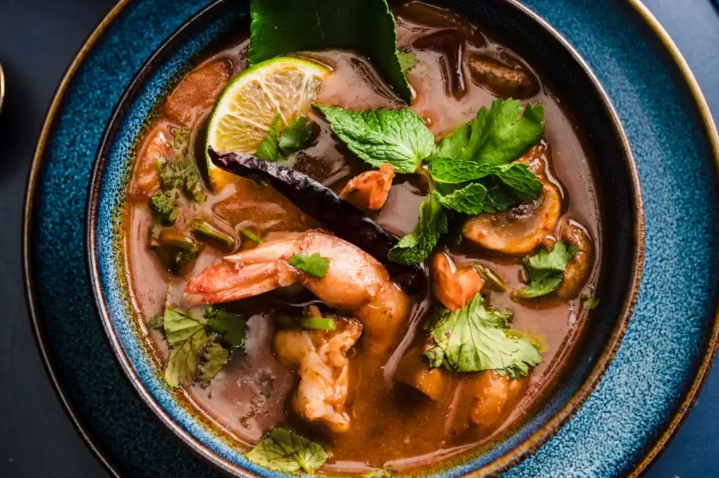 Close-up of tom yum soup with prawns, fresh herbs, lime, and Thai spices in a blue bowl at The Thai, Shawlands
