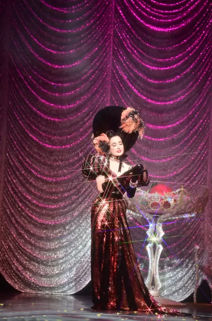 Dita Von Teese on stage during her Nocturnelle show, wearing a dramatic sequin gown and oversized hat — part of her UK tour coming to Edinburgh in 2026