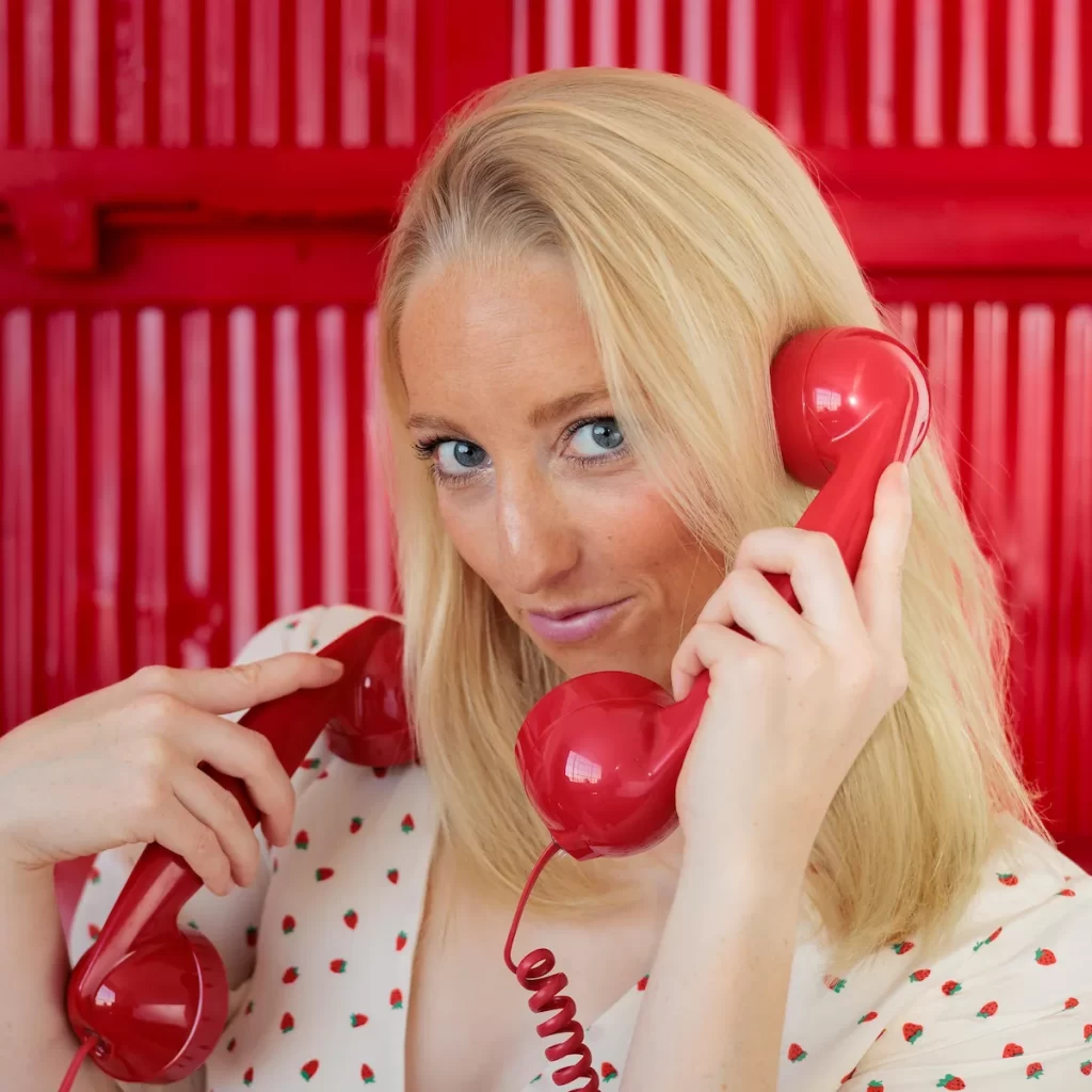 Close-up of Olivia Raine Atwood smiling with a red telephone in her hand, wearing cream