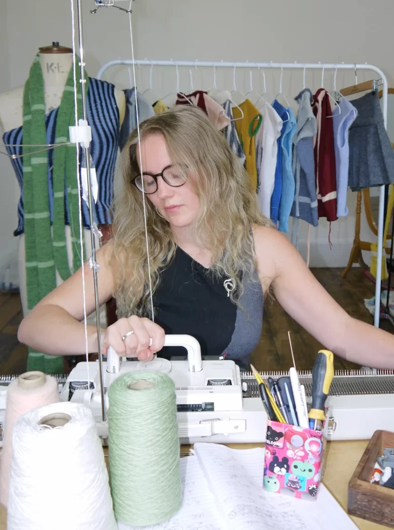 Rowan Walker, Glasgow knitwear designer, working on a manual knitting machine surrounded by yarn cones and her handmade garments.