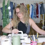 Rowan Walker, Glasgow knitwear designer, working on a manual knitting machine surrounded by yarn cones and her handmade garments.