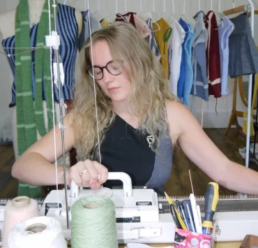 Rowan Walker, Glasgow knitwear designer, working on a manual knitting machine surrounded by yarn cones and her handmade garments.