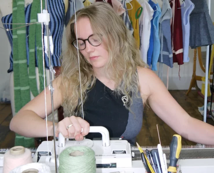Rowan Walker, Glasgow knitwear designer, working on a manual knitting machine surrounded by yarn cones and her handmade garments.
