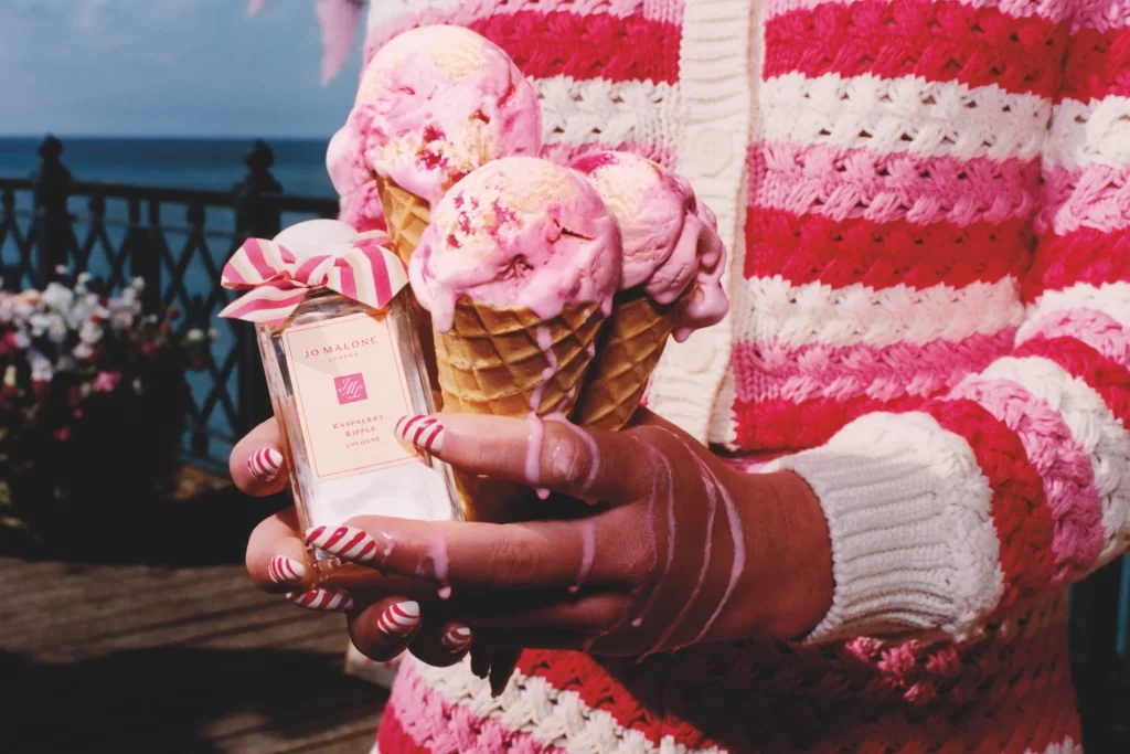 Jo Malone Raspberry Ripple cologne bottle held next to triple scoop raspberry ripple ice cream cones.