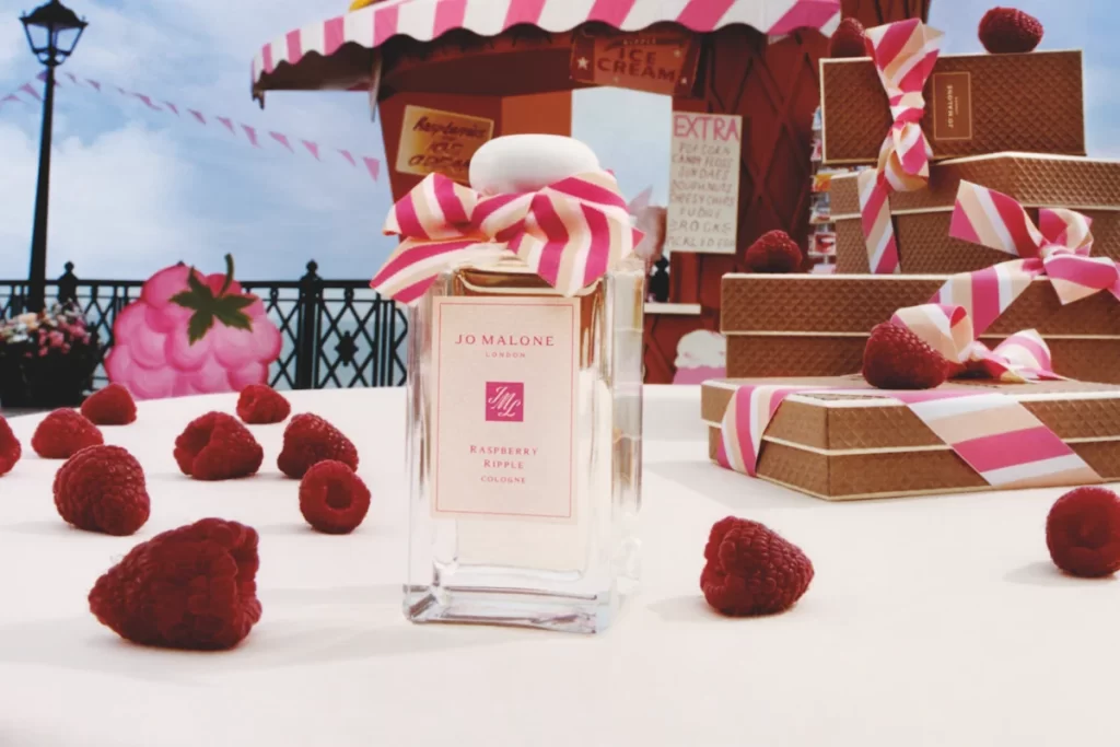 Jo Malone Raspberry Ripple cologne bottle with pink ribbon, surrounded by raspberries and waffle-textured gift boxes.