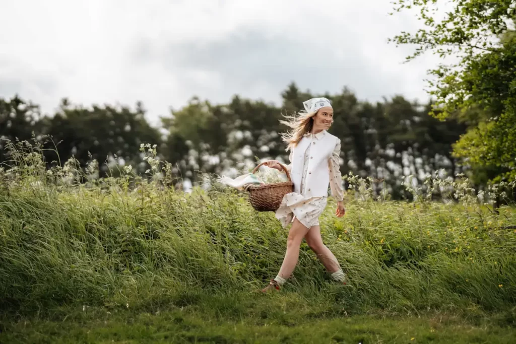 Woman in cream sleeveless dress, head-scarf and basket strolling a summer meadow.