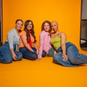 Four members of the Girl Pop! cast sitting together against a bright yellow backdrop, dressed in bold Y2K-inspired outfits.
