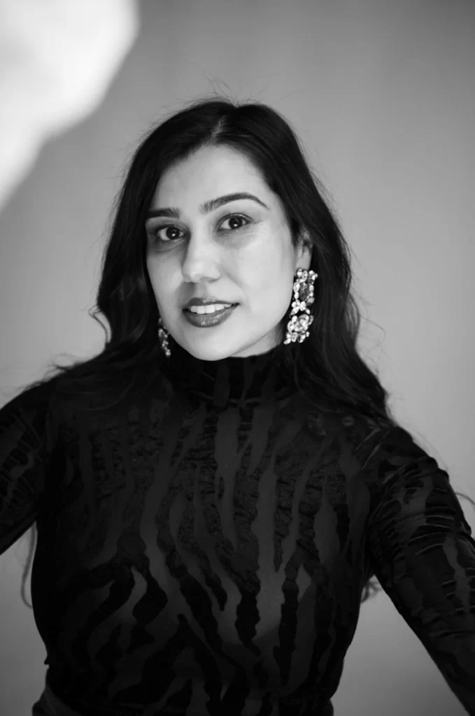 Black and white portrait of Kiran Benawra, wearing statement earrings and a patterned top, looking directly into camera.