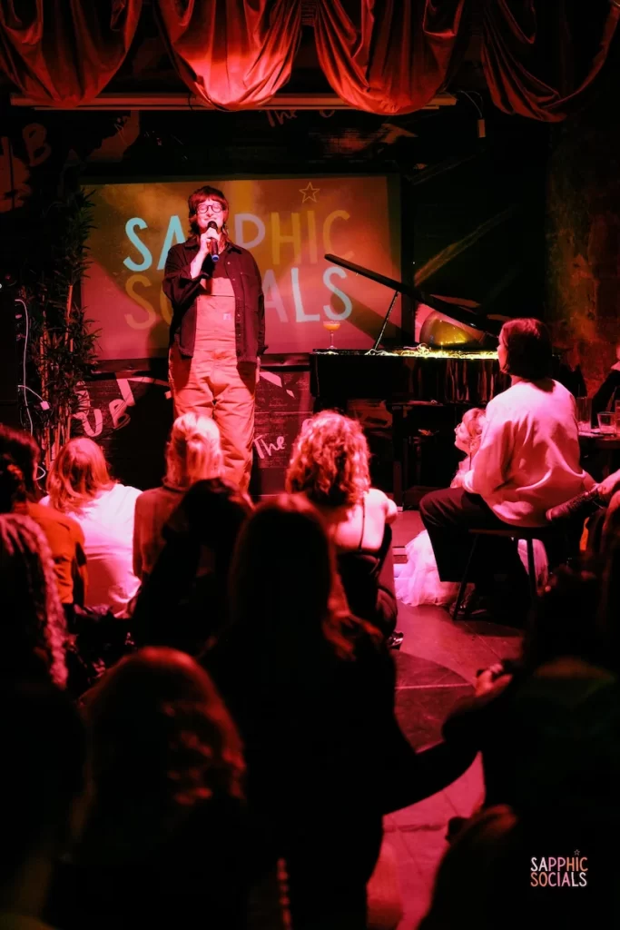 A performer speaks on stage in front of a Sapphic Space projection as the crowd watches at a Glasgow queer community event