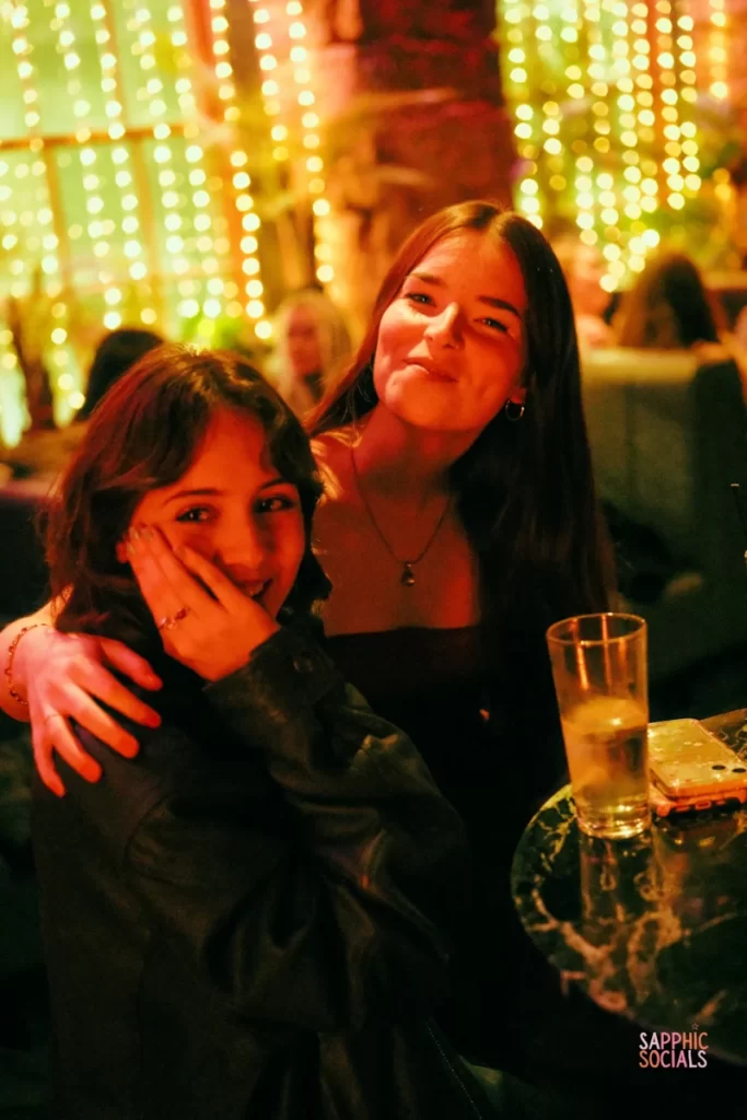 Two smiling guests at a Sapphic Space event in Glasgow lit by warm fairy lights, sitting at a cocktail table