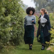 Two women in matching tartan skirts walking through a leafy garden path.