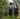 Two women in matching tartan skirts walking through a leafy garden path.