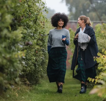 Two women in matching tartan skirts walking through a leafy garden path.
