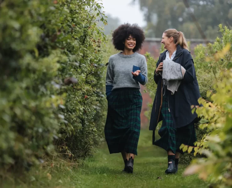 Two women in matching tartan skirts walking through a leafy garden path.
