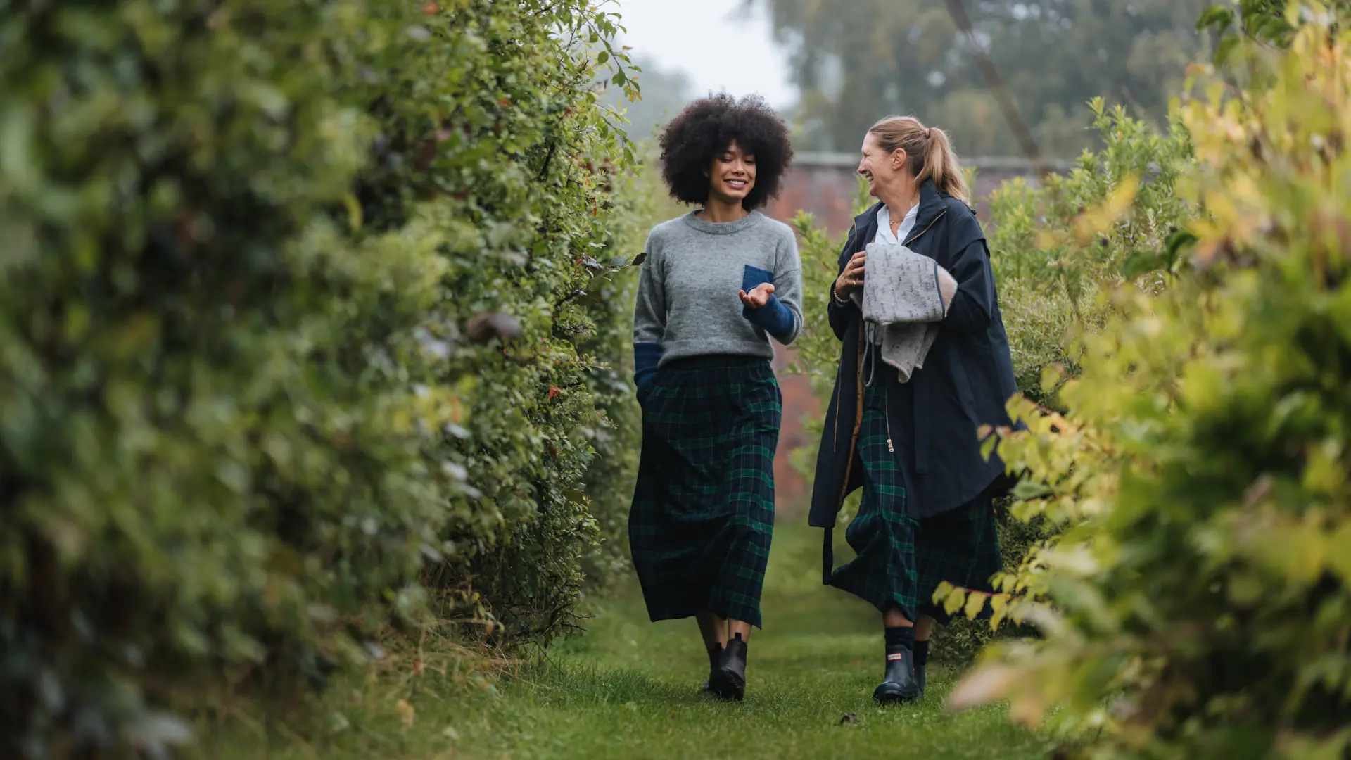 Two women in matching tartan skirts walking through a leafy garden path.