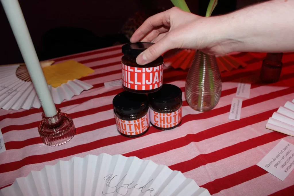 A hand placing jars of chutney and chilli jam on a red-striped table.