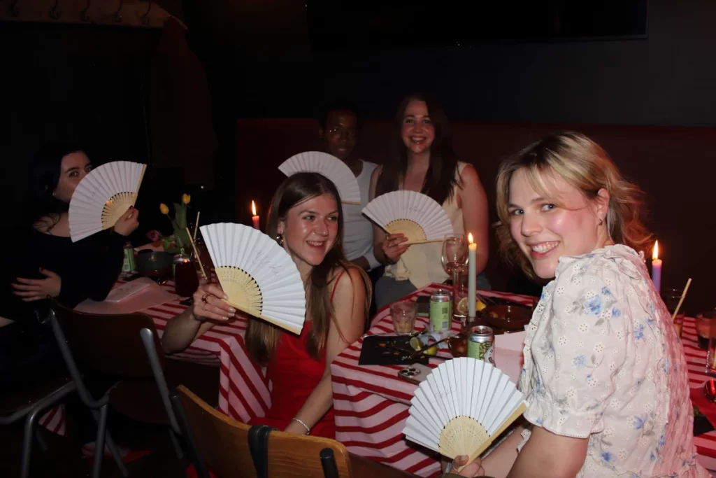 Women seated at a table holding white fans with calligraphy names.