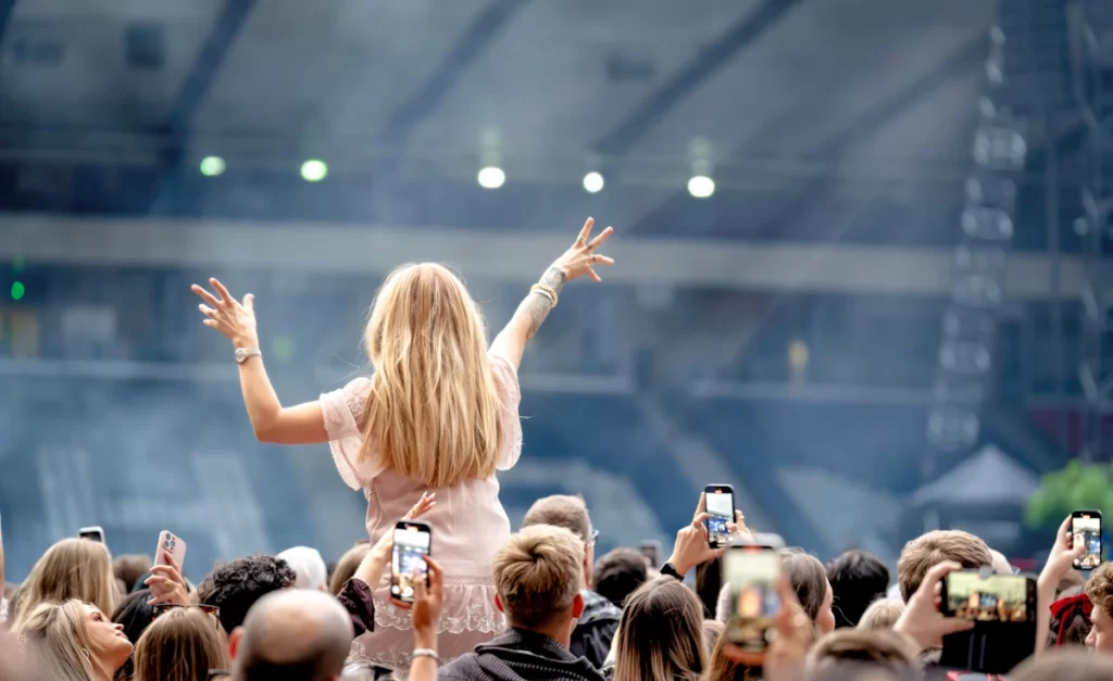 Woman on someone’s shoulders with arms raised during a live concert at Hampden Park, surrounded by a cheering crowd.