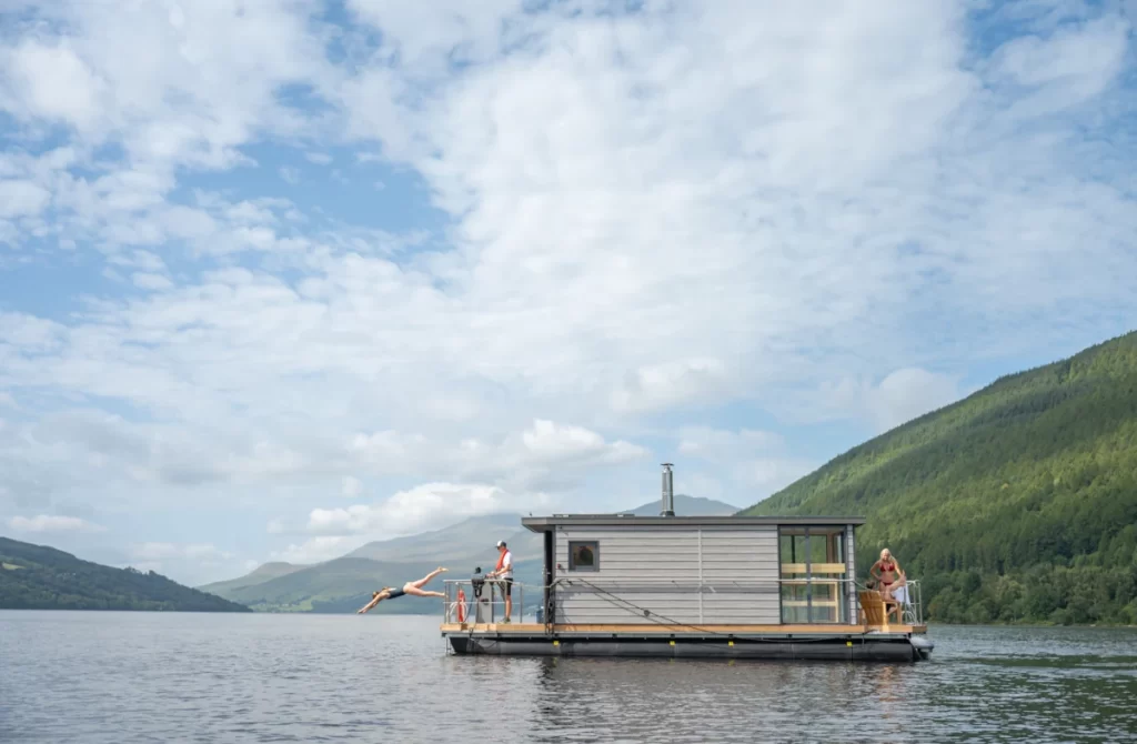 Floating sauna boat on Loch Tay with a woman diving into the water, a skipper at the helm, and another guest relaxing on deck.