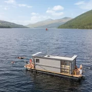 Overhead view of the Taymouth Marina HotBoat floating sauna with guests swimming in Loch Tay, surrounded by rolling hills and open water.
