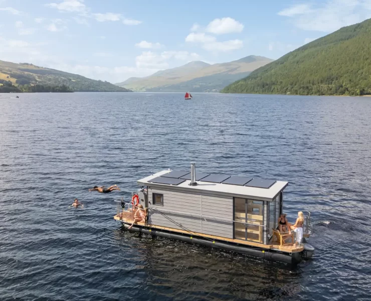 Overhead view of the Taymouth Marina HotBoat floating sauna with guests swimming in Loch Tay, surrounded by rolling hills and open water.