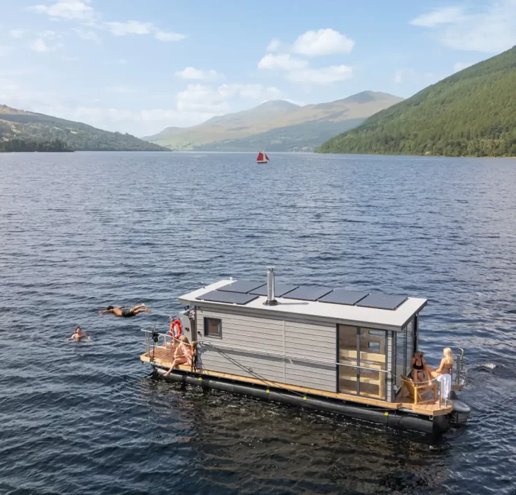Overhead view of the Taymouth Marina HotBoat floating sauna with guests swimming in Loch Tay, surrounded by rolling hills and open water.