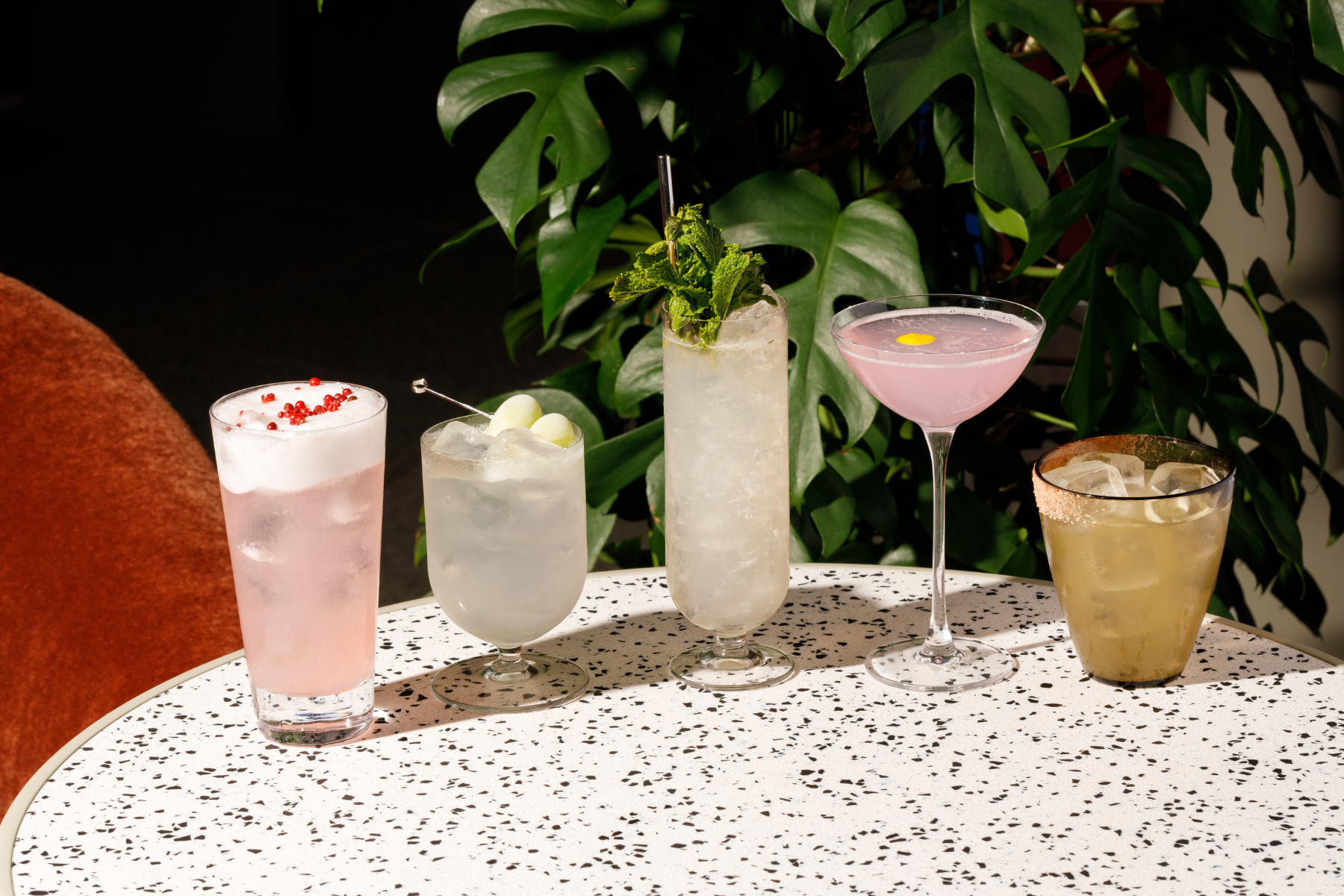 Five cocktails in different glasses on a terrazzo table, including a pink spritz, a short glass with melon garnish, a tall mojito-style drink, a pink martini and a golden cocktail rimmed with spice.