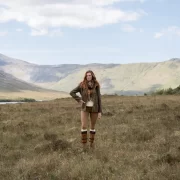 Model wearing Dubarry country boots, leggings, and a green jacket standing in a scenic Highland landscape with mountains and loch in the background.