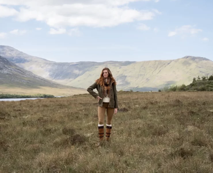 Model wearing Dubarry country boots, leggings, and a green jacket standing in a scenic Highland landscape with mountains and loch in the background.