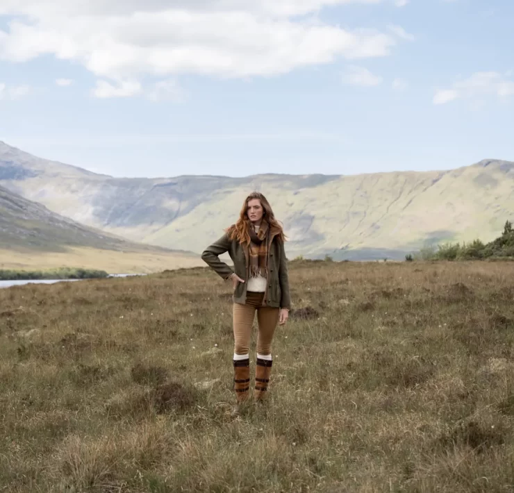 Model wearing Dubarry country boots, leggings, and a green jacket standing in a scenic Highland landscape with mountains and loch in the background.