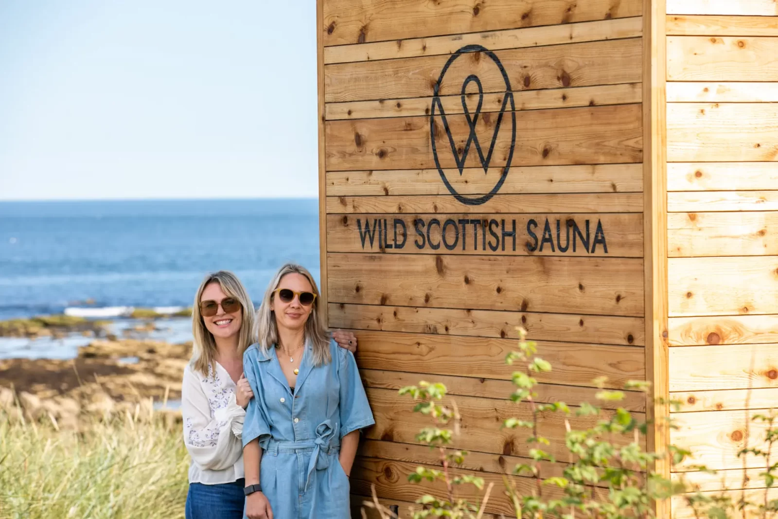Two women standing by a Wild Scottish Sauna cabin at Kingsbarns beach in Scotland.