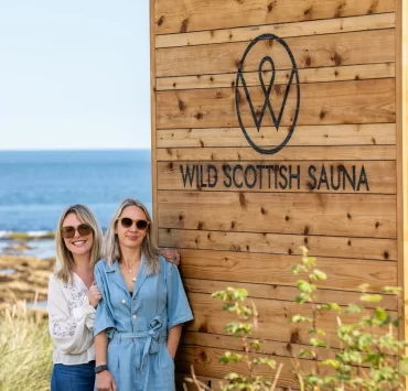 Two women standing by a Wild Scottish Sauna cabin at Kingsbarns beach in Scotland.