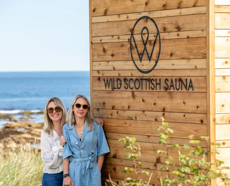 Two women standing by a Wild Scottish Sauna cabin at Kingsbarns beach in Scotland.
